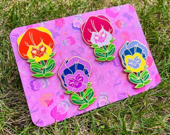 May include: Four enamel pins featuring cartoon faces on a flower design. The flowers are red, pink, purple, and blue. The pins are on a pink background with a floral pattern.