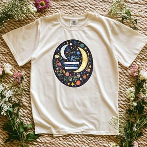 May include: White t-shirt featuring a circular graphic with a crescent moon, stars, and floral elements. The graphic includes text. The shirt is surrounded by flowers on a woven background.