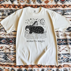 May include: Cream-colored t-shirt featuring a black and white illustration of two figures under a crescent moon and stars. The text "midwife magic" is printed below the illustration. The shirt has short sleeves and a crew neck.