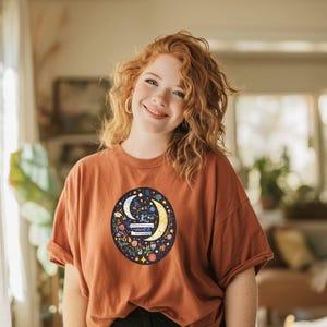 May include: Rust-colored t-shirt featuring a circular graphic with a crescent moon, stars, and floral elements. The design is centered on the shirt, creating a visually appealing focal point. The shirt has short sleeves and a relaxed fit.