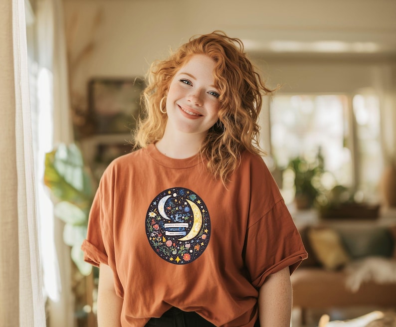 May include: Rust-colored t-shirt featuring a circular graphic with a crescent moon, stars, and floral elements. The design is centered on the shirt, creating a visually appealing focal point. The shirt has short sleeves and a relaxed fit.