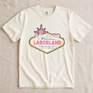 May include: A cream-colored short-sleeve t-shirt featuring a graphic design shaped like a retro sign. The text reads Welcome to Fabulous LABORLAND where the only way out is through. The design includes a pink star and yellow border accents.