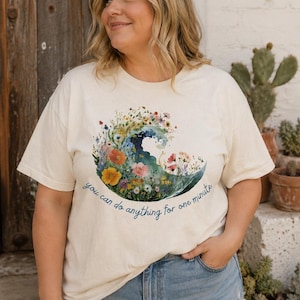 May include: A cream-colored short-sleeve t-shirt featuring a graphic of a wave composed of colorful wildflowers. Below the wave, the text reads, you can do anything for one minute in a dark blue script font. The shirt is paired with blue denim jeans.