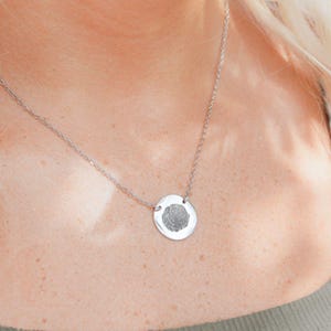 Può includere: Una collana color argento con un ciondolo circolare con un'impronta digitale. Il ciondolo è attaccato a una catena sottile. La collana è indossata intorno al collo.