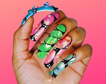 Puede incluir: Una mano que muestra uñas acrílicas largas y coloridas. Cada uña presenta un diseño único con detalles de mariposas, lunares blancos y negros y colores vibrantes como rosa, verde y azul. El texto "babynail.girl" está en la parte inferior.
