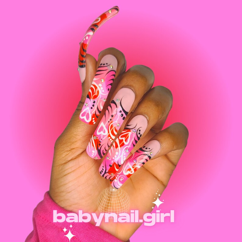 Op de afbeelding: Een hand met lange, vierkant gevormde, geverfde nagels tegen een roze achtergrond. De nagels zijn voornamelijk roze met rode en witte hartontwerpen, zwarte accenten en strass-steentjes. De tekst "babynail.girl" staat onderaan.