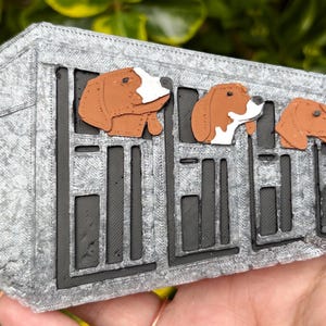 May include: A rectangular, silver-colored, textured object featuring a black grid pattern resembling a kennel. Four brown and white dog heads are positioned along the top edge, appearing to look out from the individual sections of the structure.
