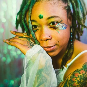 May include: A person with green and blue dreadlocks, wearing a white sheer top and a flower tattoo on their arm. They have green and blue face paint and a nose ring.