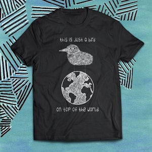 Puede incluir: Camiseta negra con un gráfico blanco de un pájaro de pie en la cima del mundo. El texto "this is just a bird on top of the world" está impreso en la camiseta.