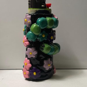 May include: A black lighter decorated with three-dimensional sculpted elements including two green caterpillars and several small flowers in shades of pink, purple, and yellow. The lighter features a metallic top with a red ignition button.