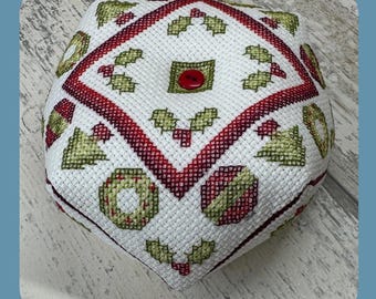 Op de afbeelding: Een wit kruissteekpatroon met een rood en groen kerstthema. Het patroon bevat hulstbladeren, ornamenten en een krans. Het patroon is gestikt op een witte stof en bevat een rode knoop in het midden.