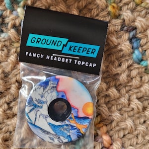 May include: A "GROUND KEEPER" fancy headset topcap in a clear plastic bag. The topcap features a mountain landscape design with blue, white, orange, and yellow colors. The topcap has a black center hole.