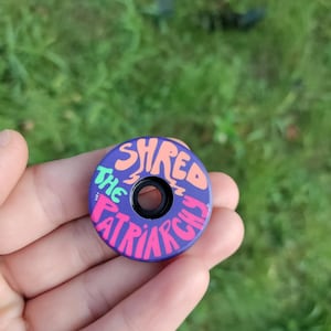 May include: A purple round bike headset cap with the words "Shred the Patriarchy" written in pink, green, and orange letters.