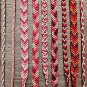 Puede incluir: Una colección de pulseras de la amistad hechas a mano en varios tonos de rojo, rosa y blanco. Las pulseras presentan diferentes patrones tejidos, incluyendo diseños de chevrón y trenzados, sobre una superficie de madera.