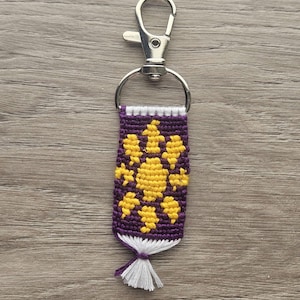 May include: A handmade keychain featuring a purple and yellow woven design with a silver clasp. The woven pattern includes yellow floral shapes on a purple background, with a white tassel at the bottom. The keychain is designed for keys or accessories.
