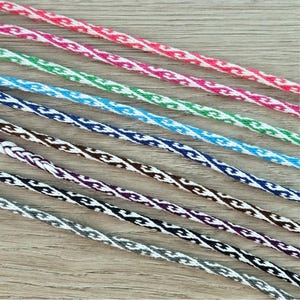 May include: A collection of eight braided cords arranged in parallel, each featuring a two-tone pattern of white intertwined with red, green, light blue, dark blue, brown, purple, black, and gray threads, creating a consistent woven texture.