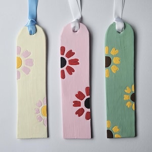 May include: Three rectangular bookmarks with rounded tops and ribbon loops. The first is pale yellow with light pink flowers, the second is light pink with red flowers, and the third is sage green with yellow flowers, all featuring dark centers.