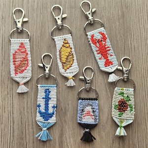 May include: A collection of handmade keychains with various nautical and animal designs. Each keychain features a woven design with a silver clasp. Designs include a seashell, lobster, anchor, penguin, and turtle. The keychains are in shades of red, blue, yellow, and green.