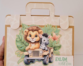 May include: A brown cardboard box shaped like a suitcase with a handle. The box is decorated with a jungle scene featuring a lion and a zebra riding in a jeep. The text "Eilam Wild One" is on the front of the box.