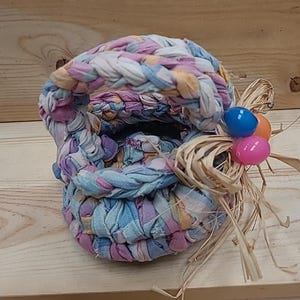 May include: A handmade Easter basket crafted from colorful fabric strips in shades of blue, purple, and yellow. The basket features a braided handle and is adorned with decorative straw and three plastic Easter eggs.