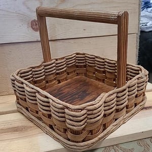 May include: A square woven basket with a wooden handle. The basket is made of natural materials and has a rustic look.