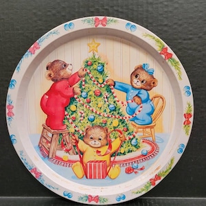 May include: A vintage metal tray with a Christmas scene featuring three teddy bears decorating a Christmas tree. The bears are wearing pajamas and are holding ornaments and ribbon. The tray has a white background with a woodgrain pattern.