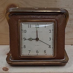 May include: A brown leather-covered alarm clock with a white face and gold trim. The clock face has black hands and black numbers. The clock face also has the text "Phinney Walker" and "Alarm".