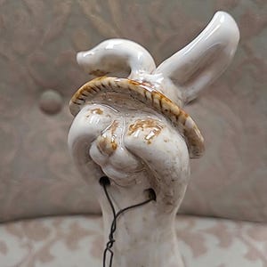 May include: A white ceramic rabbit figurine with a brown hat and a wire tail. The rabbit has a whimsical expression and is glazed with a glossy finish.