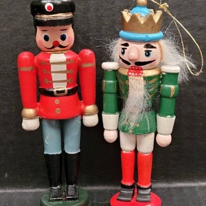 May include: Two wooden nutcracker ornaments, one in red and blue and the other in green and white. Both have painted faces and white beards.