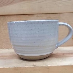 May include: A ceramic mug with a speckled light gray finish and a series of horizontal ridges along the body. The mug features a curved handle on the right side and a smooth, unglazed base that reveals the natural brown clay texture of the material.