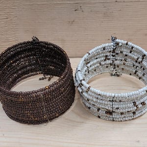 May include: Two beaded bracelets, one brown and one white, with a woven design. The bracelets are made of small beads and are stacked on top of each other.