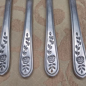 May include: Four silver-toned metal cutlery handles arranged vertically, each featuring a recessed floral motif at the base. The design consists of a single rose bloom at the bottom with a stem extending upward, flanked by small leaves on both sides.