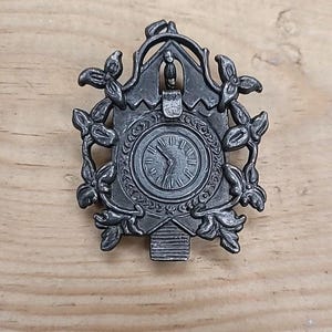 May include: A silver-toned metal cuckoo clock pin. The pin features a clock face with Roman numerals, surrounded by floral and leaf designs. The clock has a decorative top and a pendulum at the bottom.