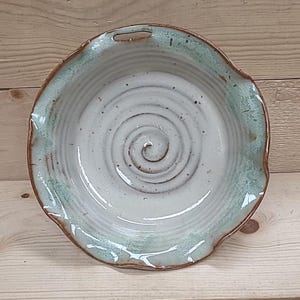 May include: Handmade ceramic bowl with a spiral design in the center. The bowl has a light blue-green rim and a brown edge. The interior is a light cream color with a spiral pattern. The bowl has a small handle on the rim.