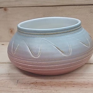 May include: A ceramic bowl with a rounded shape and a wide opening. The bowl features a gradient color scheme, transitioning from a pinkish-red at the base to a light blue at the rim. Decorative wave-like patterns are carved into the exterior.