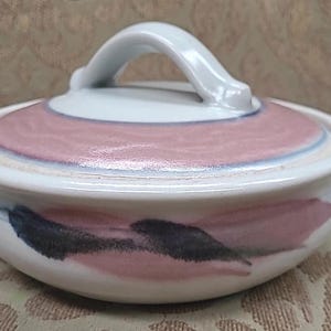 May include: A ceramic lidded bowl with a white base and lid. The lid features a pink and blue design, and a white handle. The bowl's exterior is decorated with pink and black brushstrokes.