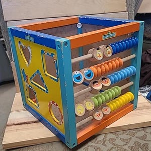 May include: A wooden Sesame Street activity cube with a colorful abacus and shape sorter. The cube features Sesame Street characters like Big Bird, Cookie Monster, and Elmo.