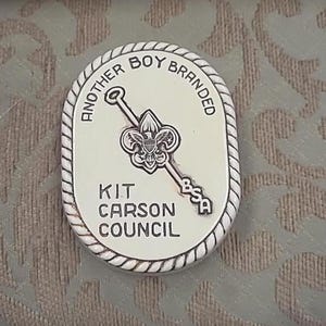 May include: Oval-shaped silver-colored pin with the text "ANOTHER BOY BRANDED" around the edge. The pin features a Boy Scouts of America emblem and the words "KIT CARSON COUNCIL".