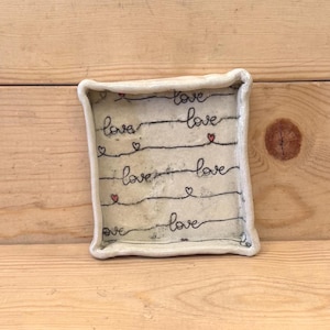 May include: A square, off-white ceramic trinket dish with raised, wavy edges. The surface features horizontal black lines with the handwritten word love repeated multiple times, interspersed with small, hand-painted red hearts.
