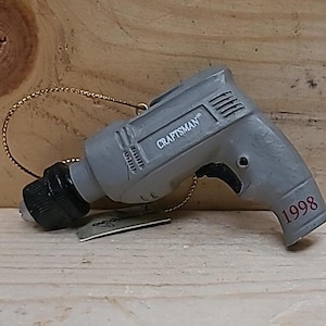 May include: A gray and black Craftsman drill ornament with the year 1998 printed on the side. The ornament is made of plastic and has a gold string attached to it.