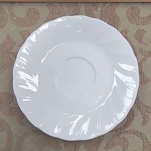May include: A white saucer with a gently scalloped edge. The center of the saucer has a recessed area for a teacup. The design is simple and elegant, suitable for a formal tea setting.