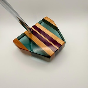 May include: A decorative putter head with a clear shaft. The head is made of wood and resin, featuring stripes of natural wood, purple, and teal. The putter is angled, suggesting it is in use.