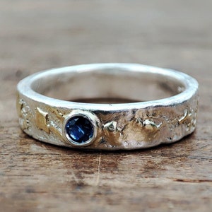 May include: A silver ring with a textured surface and a small blue gemstone set in the center. The ring has a band of gold around the edge.