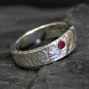May include: A silver ring with a textured finish and a single red gemstone set in the band.