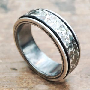 May include: A silver ring with a textured band and a black border. The ring has a hammered metal design, giving it a rustic and unique appearance. The ring is a piece of jewelry.