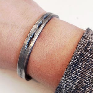 May include: A silver-toned metal cuff bracelet with a textured, distressed design. The bracelet has a rugged, industrial aesthetic, with irregular cut-outs along its surface. The cuff is worn on a wrist, showcasing its unique, handcrafted appearance.