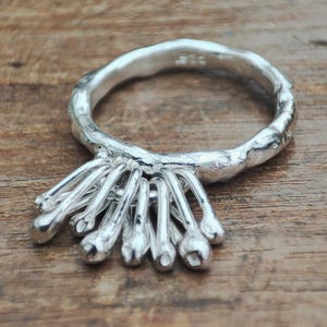 May include: A silver ring with a textured band and a cluster of teardrop-shaped pendants. The ring is made of a shiny, metallic material and sits on a wooden surface. The ring's design is organic and modern.