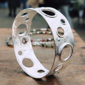 May include: A silver-colored bracelet with a textured surface and multiple oval and circular cut-out holes. The bracelet is displayed on a wooden surface, with a blurred background.