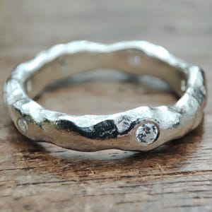 Può includere: Un anello d'argento con un design strutturato e organico. L'anello presenta diverse piccole pietre preziose chiare incastonate nella fascia. L'anello è appoggiato su una superficie di legno.