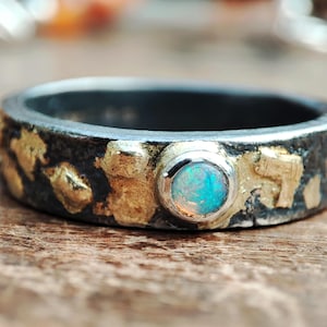 May include: A silver ring with a central, round, iridescent opal stone. The band has a dark, textured finish with gold-colored accents. The opal displays shades of blue and green.